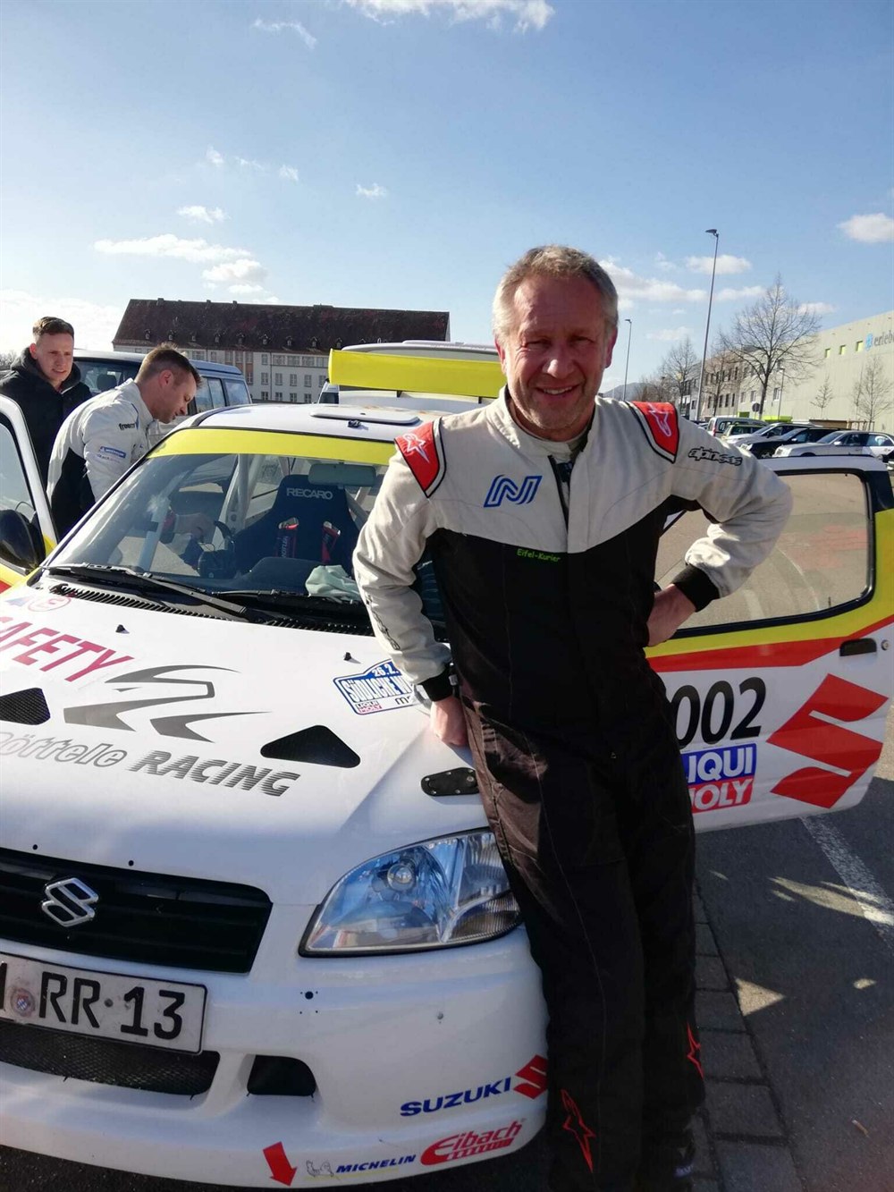 Niki Schelle im Rennanzug mit Eifel-Kurier Schriftzug vor seinem Suzuki Rallyefahrzeug